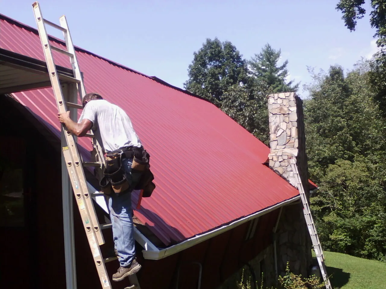 Roofer on a ladder beside a red metal roof and stone chimney — Soffit & Fascia Repair by Roofing Brothers in Watervliet